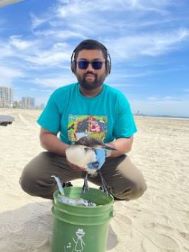 Bangladeshian man at a beach clean up. He is wearing headphone, teal shirt and brown pants and is holding a bucket