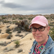 Human standing in a desert. They have a pink baseball cap on and cat eye glasses