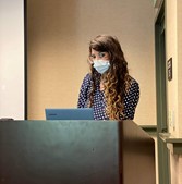 Female grad student lecturing. She is wearing a face mask and has long brown curly hair. 