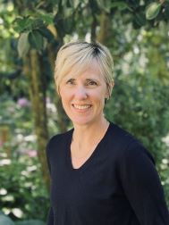 Short hair blonde woman with a black vee neck tee on. Posing in a garden. Headshot