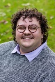 Dark curly haired person posing for a headshot. They have heart shaped glasses on. 