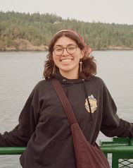Woman posing on a boat. She has her hair in a bandana and is wearing a black sweatshirt. 