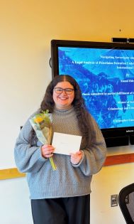 Student with long curly dark hair holding a bouquet of flower and an envelope. It looks like they are getting an award