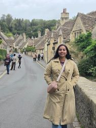 Student wearing a tan trenchcoat and a pink sling purse. She has long black hair and looks to be in a small village in England.