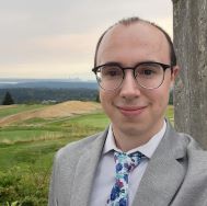Grad student posing in front of a large green field. They are wearing a grey suit and tie and they have short hair and glasses. 