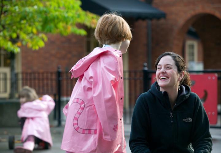 A young child in a pink raincoat has her back turned to us as she is facing a smiling brown-haired adult woman.