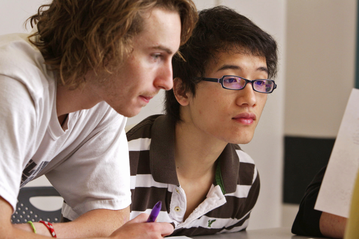 Two students, sit should to shoulder looking at something ahead. One student has short brown hair and is wearing glasses and black and white striped shirt. The other student has light brown hair and white T-shirt