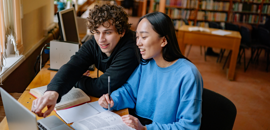 Two smiling students sit at a wooden desk in a library, looking together at a laptop screen. The student on the left, wearing a black hoodie, points to the laptop screen, while the student on the right, wearing a blue sweatshirt, holds a pen over an open notebook. Several open textbooks rest on the desk in front of them.
