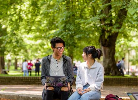 Two students sitting in front of many leafy green trees. One has a laptop.
