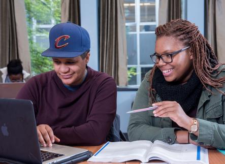 Two students sitting at a table with a laptop and book in front of them.
