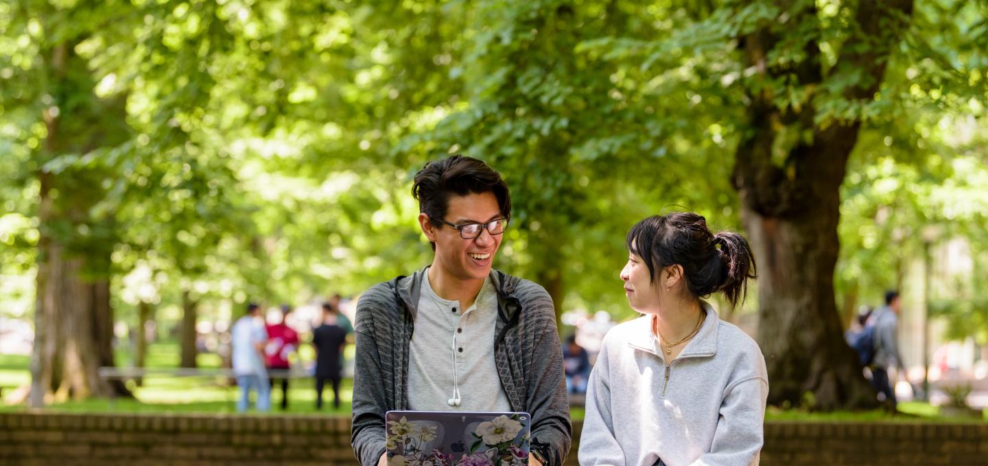 Two students sitting in front of many leafy green trees. One has a laptop.