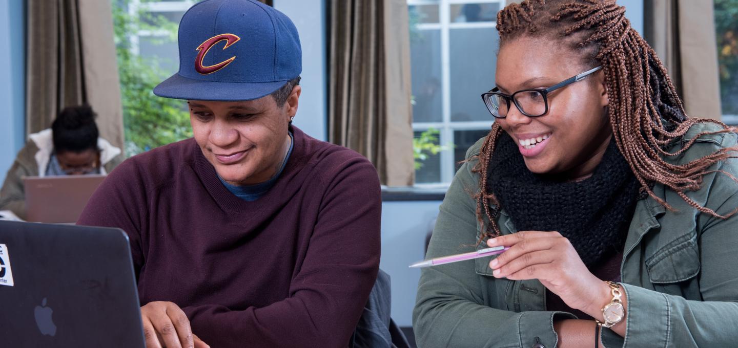 Two students sitting at a table with a laptop and book in front of them.