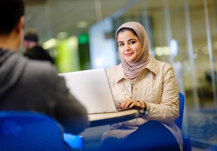 Photo looking over someone's shoulder at a student sitting at a desk with her open laptop smiling at the camera 