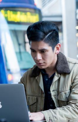 Photo of a student sitting outside Ben and Jerry's looking intently at his laptop with the streetcar in the background