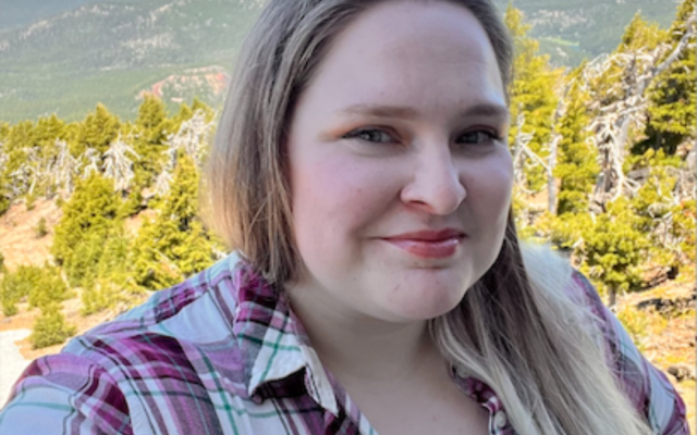 Female-identified person with long, blonde hair wearing a plaid shirt with mountains in the backround.
