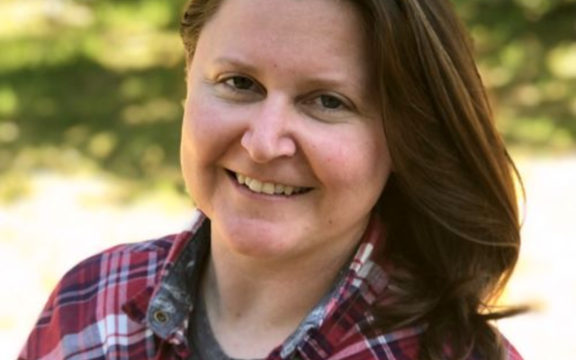 Female-identified person with medium-length brown hair, wearing a plaid shirt with trees in the background.
