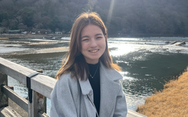 Female-identified person with long, brown hair wearing a grey coat with a lake and mountain in the background.