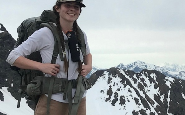 Female-identified person with long, brown hair wearing a white shirt, brown pants, and a hiking bag with mountains in the background.