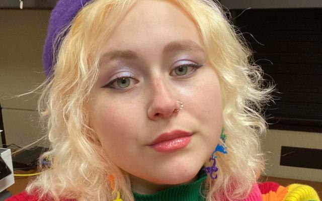 Person with short blonde hair wearing a rainbow sweater and purple hat.