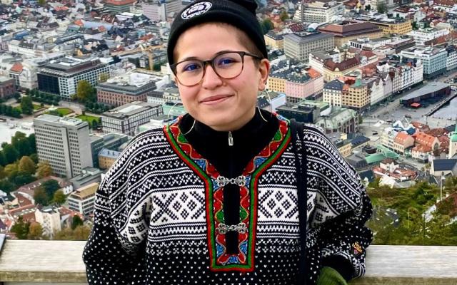 Person with short brown hair and glasses, wearing a black beanie and sweater, in front of a city skyline.