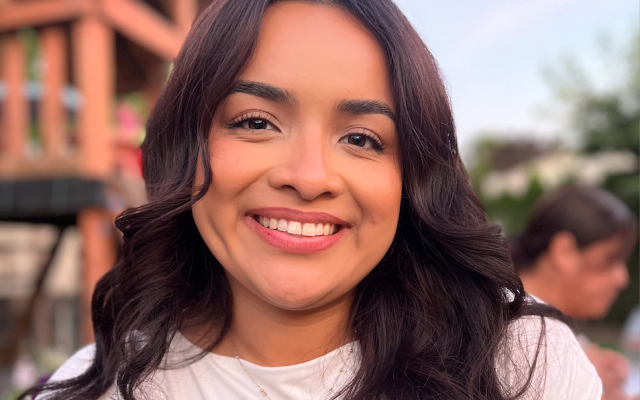 Female-identified person with long, brown hair wearing a white shirt, with a house and sky in the background.