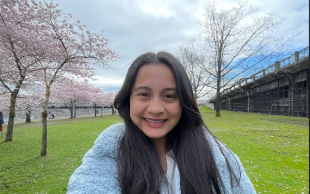 Female-identified person with long, black hair wearing a blue jacket, with trees and grass in the background.