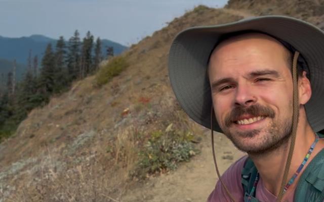 Male-identified person with brown mustache wearing a purple shirt and grey hat, with mountains in the background.