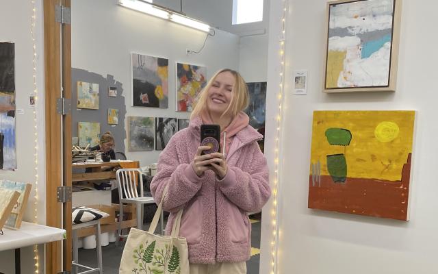 Person with short blonde hair wearing a pink sweater and cream pants, with an art studio in the background.