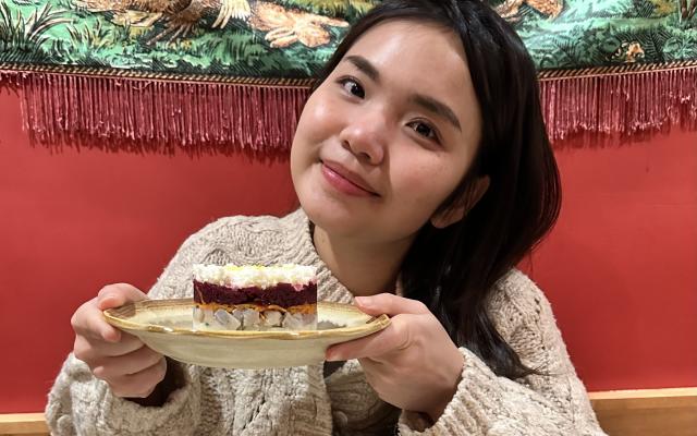 Female-identified person with long black hair, wearing a beige sweater and holding a plate with a piece of cake..
