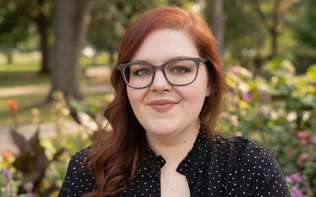 Female-identified person with long, red hair and glasses, wearing a black and white spotted top, with trees in the background.