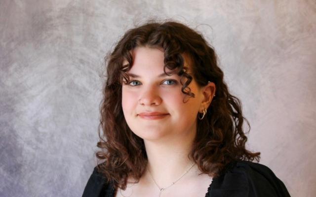 Female-identified person with medium-length curly brown hair, wearing a black shirt with a tattoo on her arm.