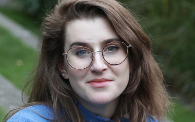 Female-identified person with long, brown hair and glasses, wearing a blue sweater, with greenery in the background.