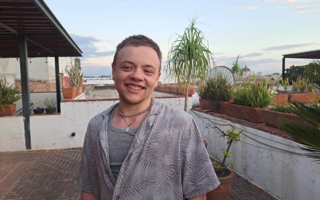 Male-identified person with short brown hair, wearing a grey shirt and necklace, with plants on a patio in the background.