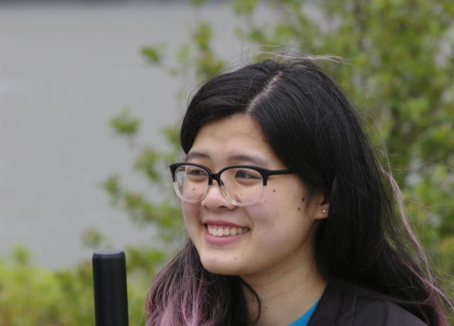 A student with black and pink hair wearing glasses looks off to the left of the camera while smiling and holding the end of a shovel