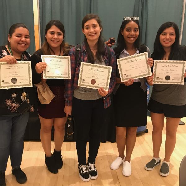 Five students stand facing the camera holding up certificates of service recognition