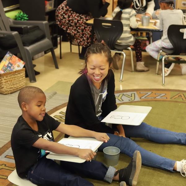 A student volunteer sits on the ground next to a kindergartener at Alder College Day