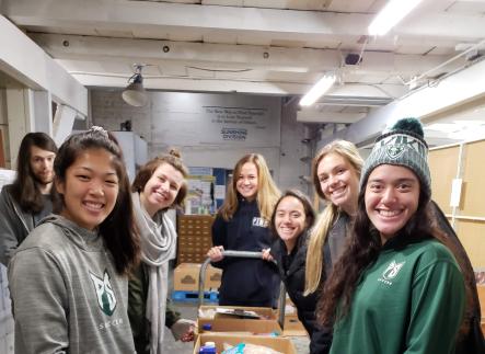 Smiling students stand around boxes of food they have packed for Sunshine Division food pantry