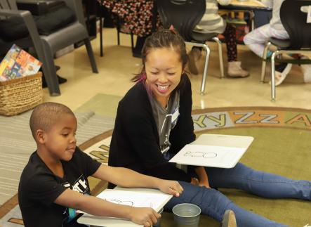 A student volunteer sits on the ground next to a kindergartener at Alder College Day