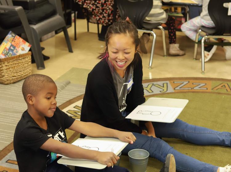 A student volunteer sits on the ground next to a kindergartener at Alder College Day