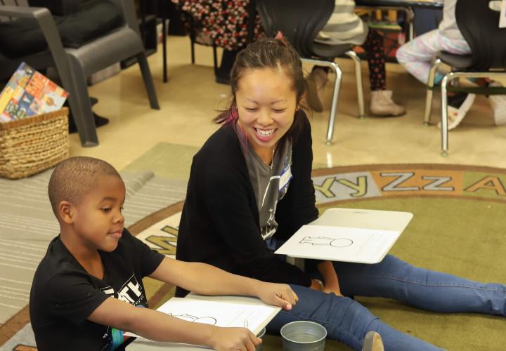 A student volunteer sits on the ground next to a kindergartener at Alder College Day