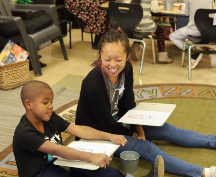 A student volunteer sits on the ground next to a kindergartener at Alder College Day