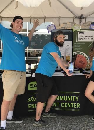 Three students wearing light blue shirts stand in front of a tabling table. One has arms raised, one holds a water bottle, and one has an arm on their hip.