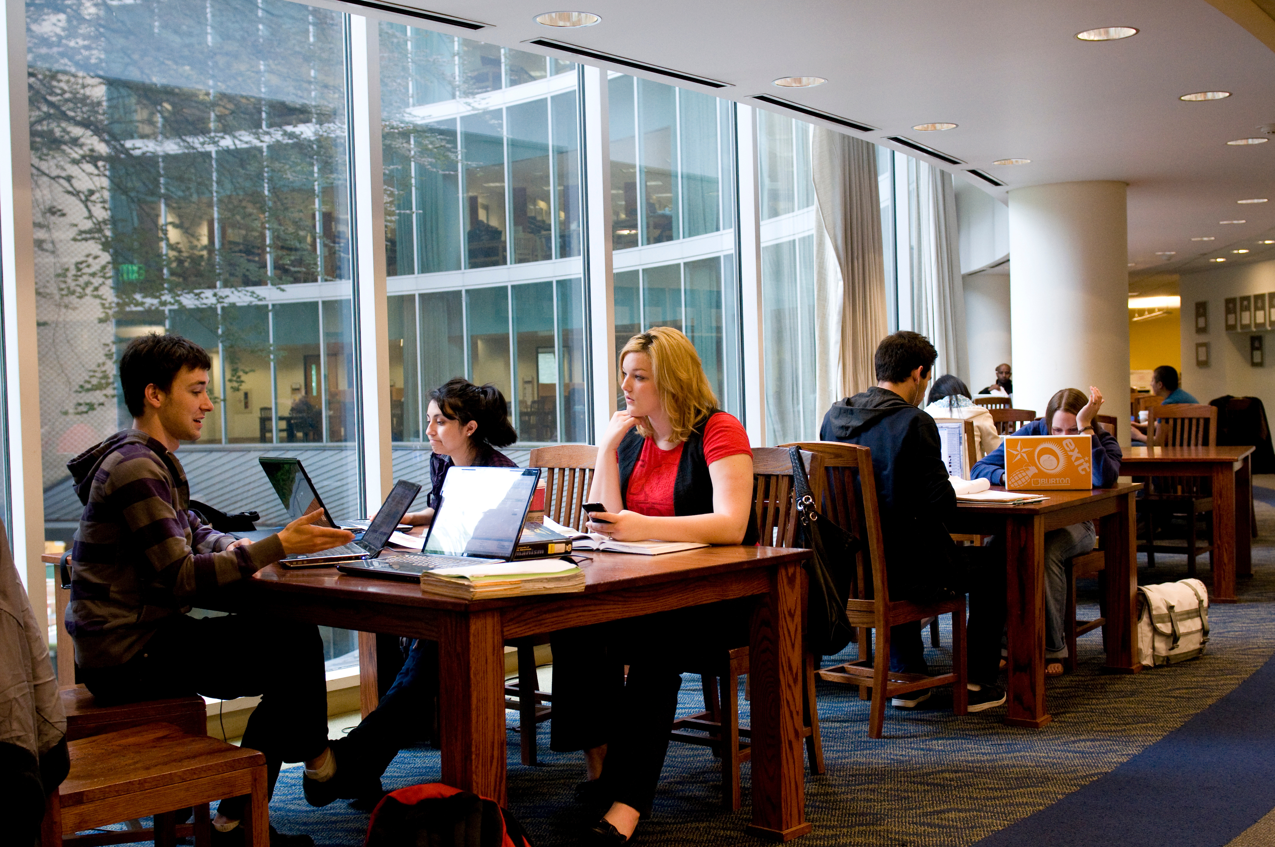 several students sitting in chairs at tables inside of library on their computers 