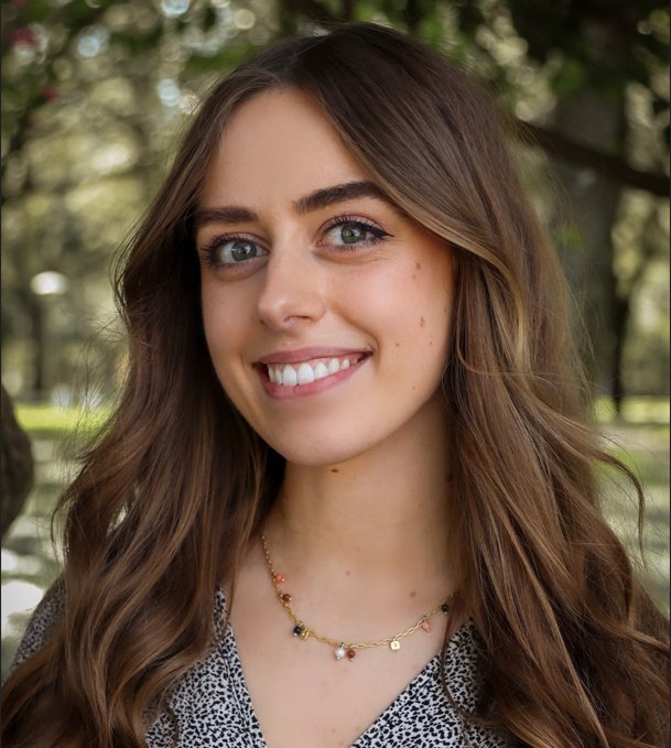 headshot of Brenna, she is facing the camera and smiling with a city park in the background. She has long brown hair, is wearing a black and white shirt.