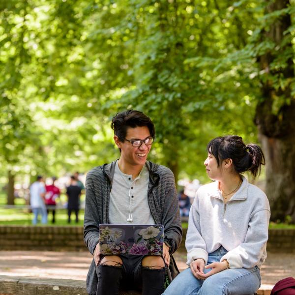 two students sitting outside on bench holding laptop and talking