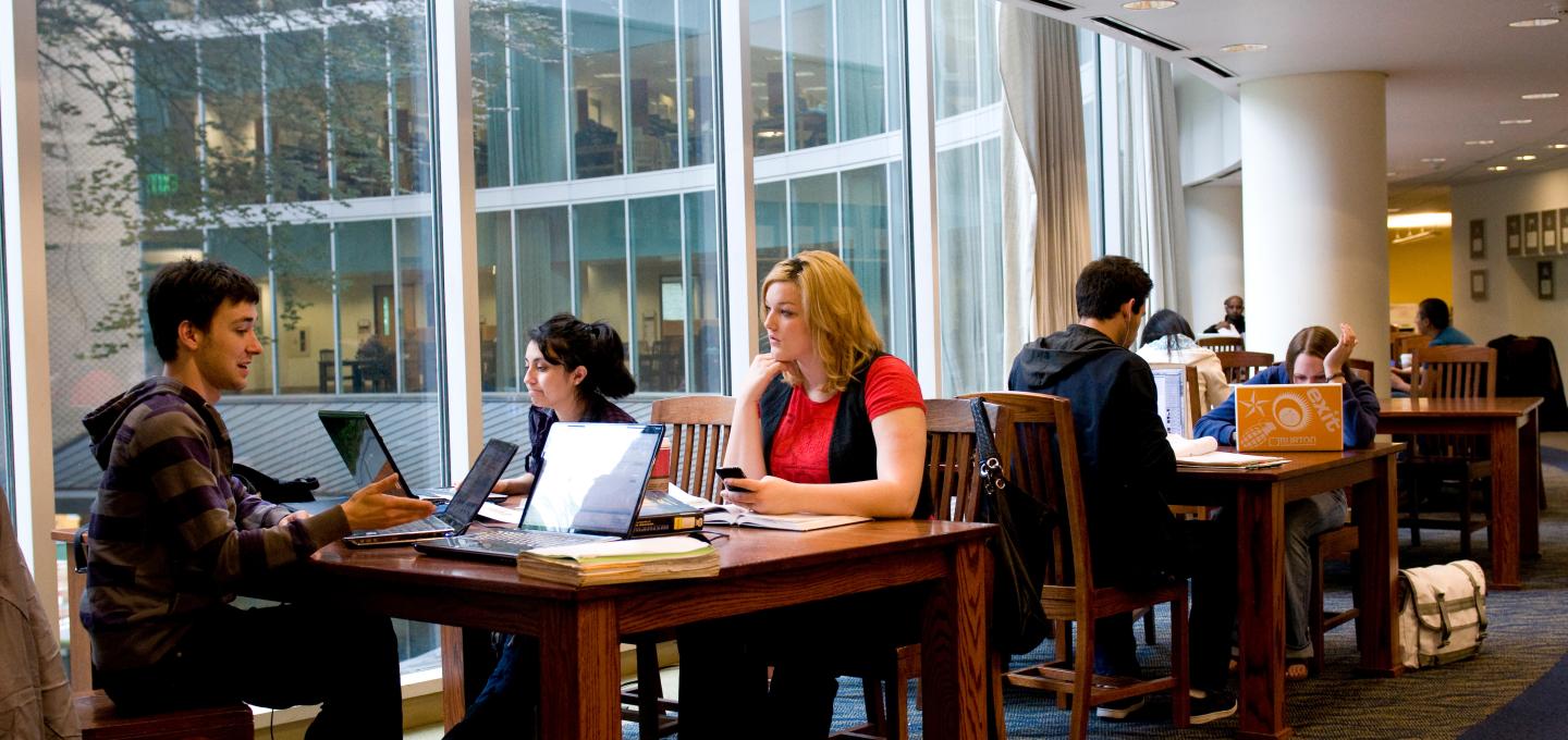 several students sitting in chairs at tables inside of library on their computers 