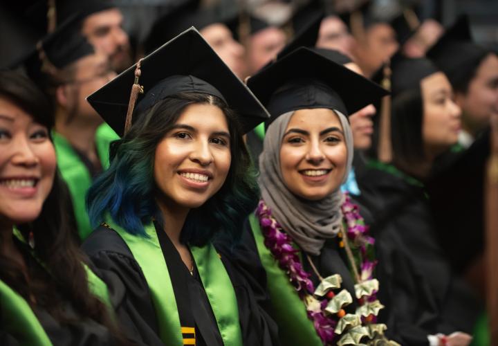 Two students at graduation, proud of their accomplishment. They are both wearing caps and gowns, a green PSU stole, the student farther from the camera is wearing a flower lei
