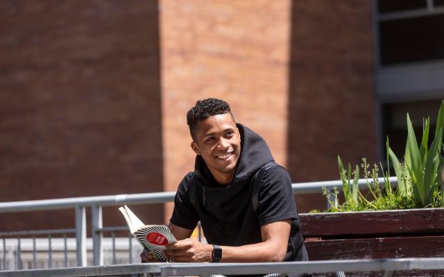 student standing on rooftop with book smiling