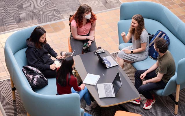 five students talking around a table with laptops and notebooks
