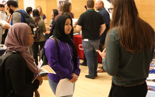 two students listening attentively to a staff member standing at a resource table. There are more tables and people in the background. 